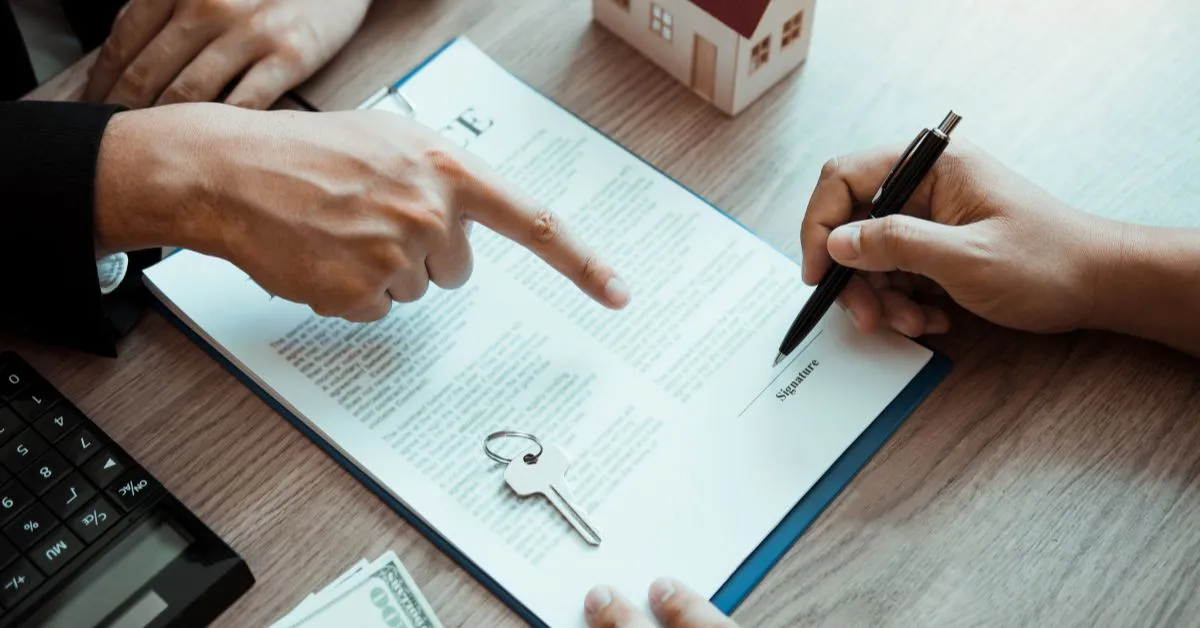 A hand signing a contract with a nearby key, calculator, and dollars