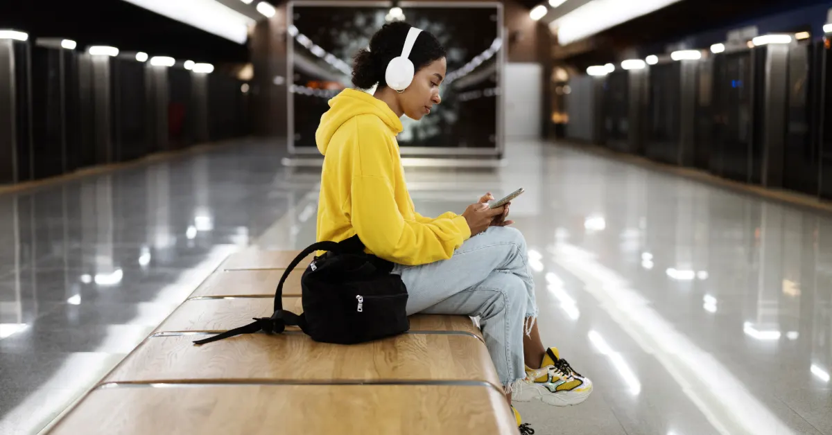 Woman in yellow hoodie with headphones using phone in subway station.