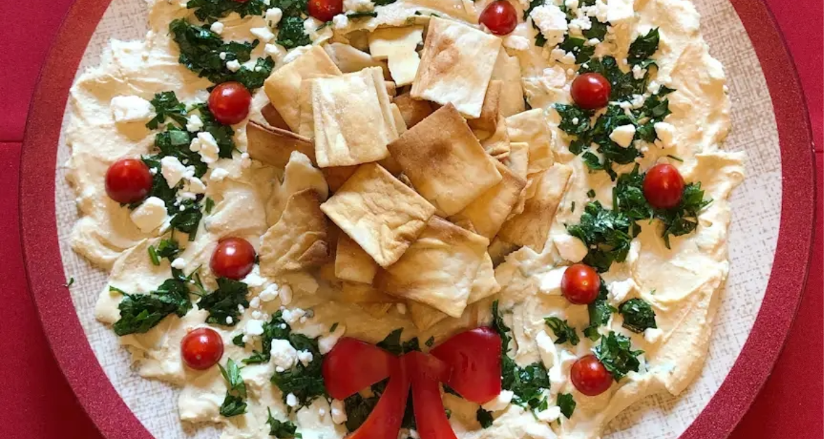 Festive hummus platter decorated with cherry tomatoes, parsley, feta, and pita chips, styled like a Christmas wreath.