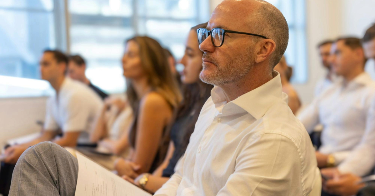 A man wearing glasses sits in front of an audience during a real estate auction, facilitating the event