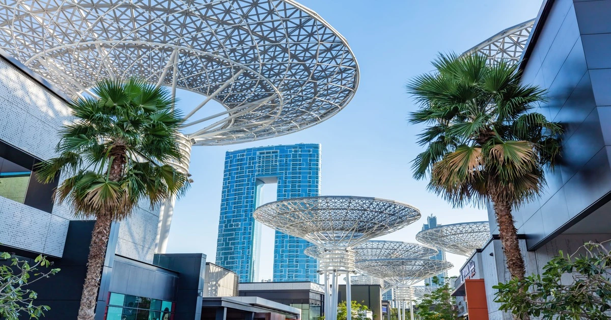 Outdoor view of a modern city walkway featuring geometric metal shade structures, tall palm trees, and a distinctive blue high-rise building.