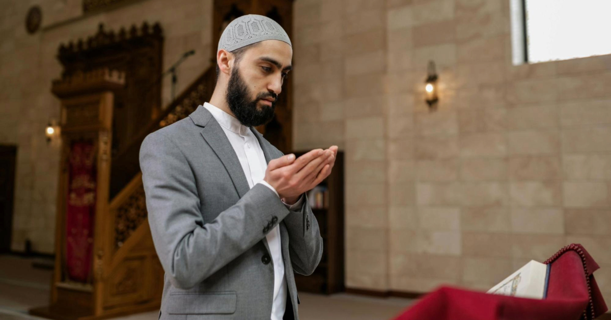 Muslim Man in a Grey Suit and Kufi Praying with Open Palms Inside a Mosque