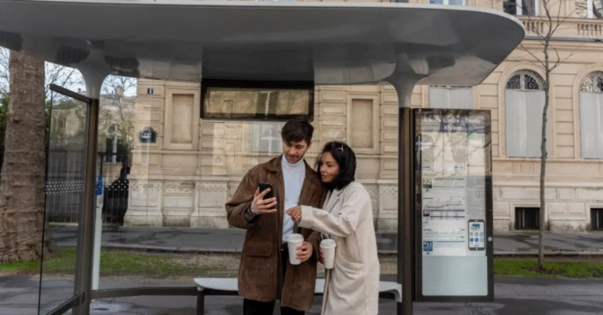 Commuters browsing their phone while waiting at a shaded bus stop