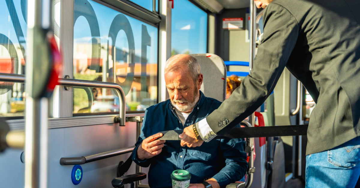 Man helping a senior in a wheelchair securing a safety belt on a bus