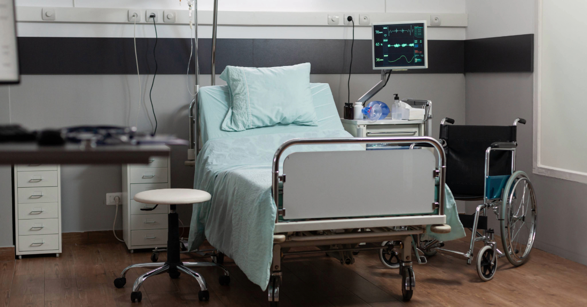 An empty hospital bed in a clean, well-lit hospital room, neatly made with white linens, ready for a patient.