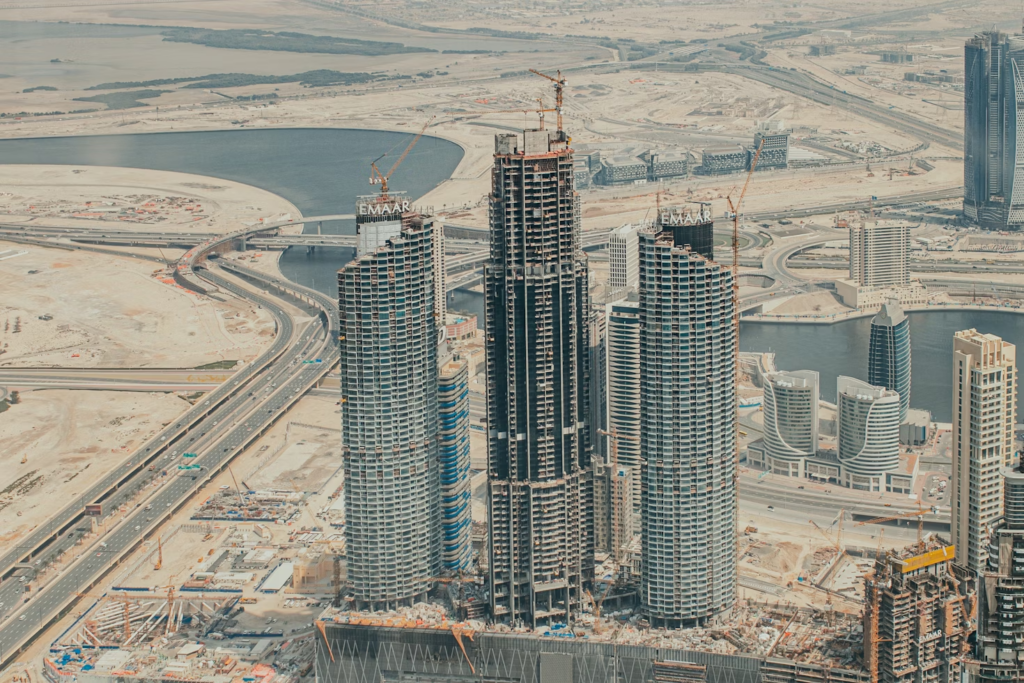 High-rise buildings under construction in Dubai with cranes atop the structures, surrounded by highways and water bodies, illustrating the city's ongoing development and urban expansion.