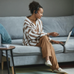 A professional woman sitting comfortably on a sofa, using a laptop and wearing a headset, engaging in a virtual meeting or sales presentation.