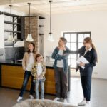 Real estate agent giving a tour of a modern kitchen to a family, explaining the features and benefits to the parents and their child. The agent is pointing to the lights and engaging with the clients, demonstrating a professional and informative approach to building relationships with homeowners and developers.