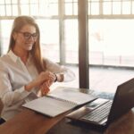 A smiling real estate professional looking at her wristwatch while working on a laptop, illustrating effective time management in a busy workspace.