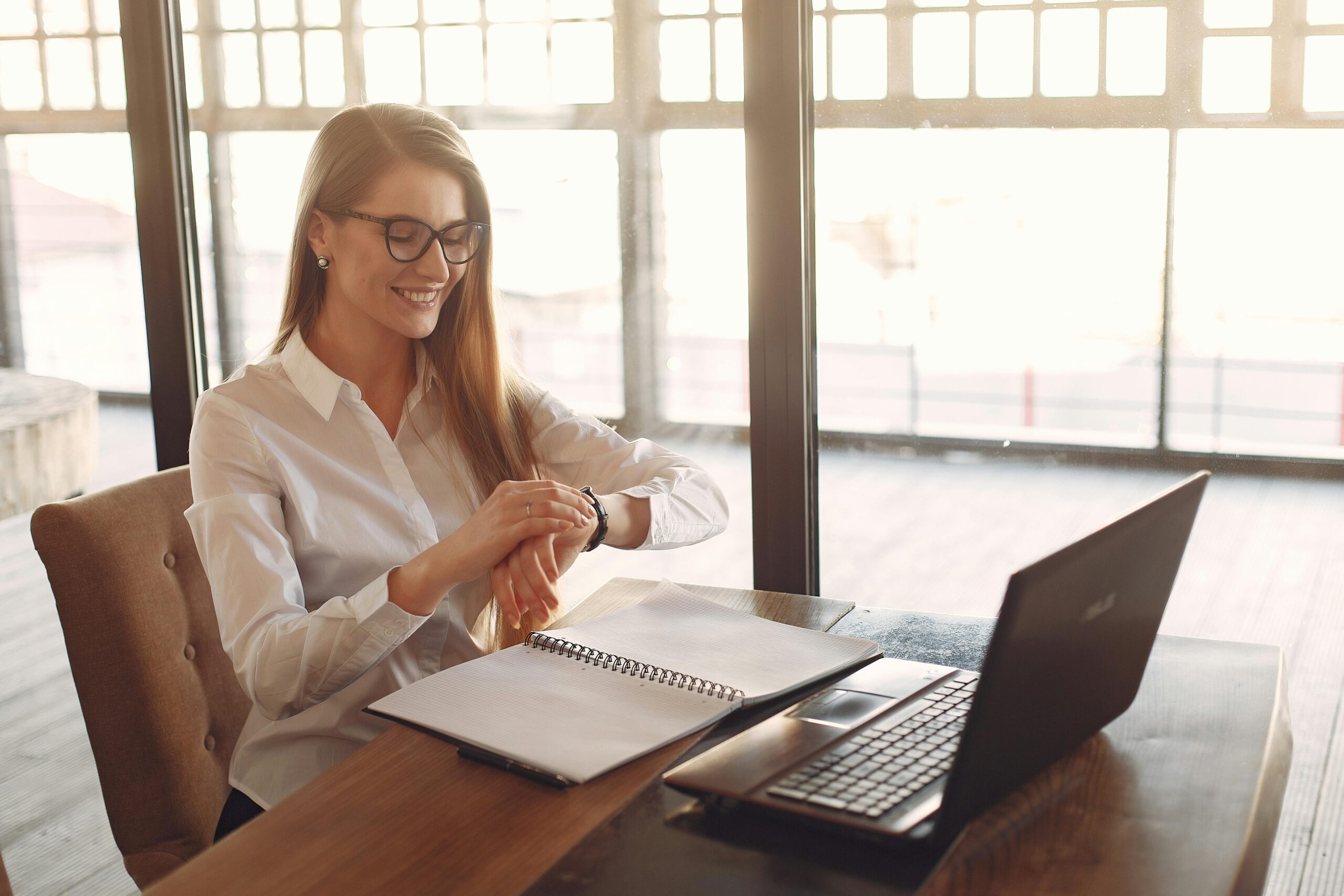 A smiling real estate professional looking at her wristwatch while working on a laptop, illustrating effective time management in a busy workspace.