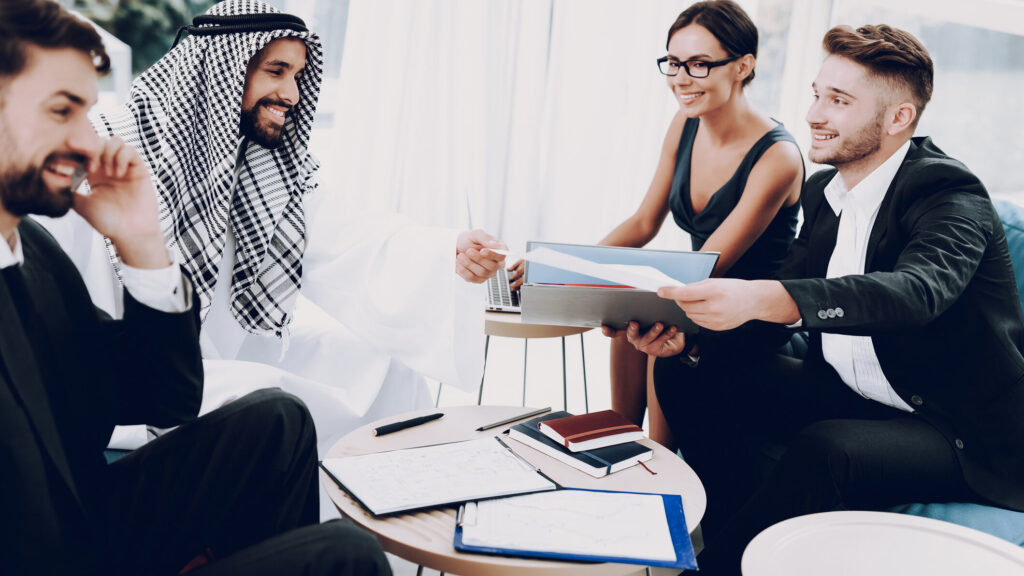 Group of professionally dressed businesspeople, including a man in traditional Middle Eastern attire, gathered around a table in a modern office setting, smiling and reviewing documents together.