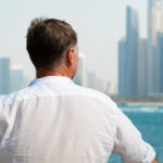 Man in a white shirt standing at a waterfront, looking out over the Dubai Marina skyline on a hazy day.