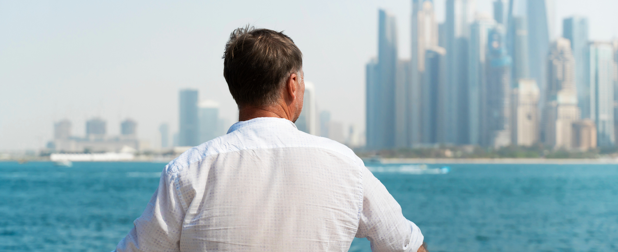 Man in a white shirt standing at a waterfront, looking out over the Dubai Marina skyline on a hazy day.