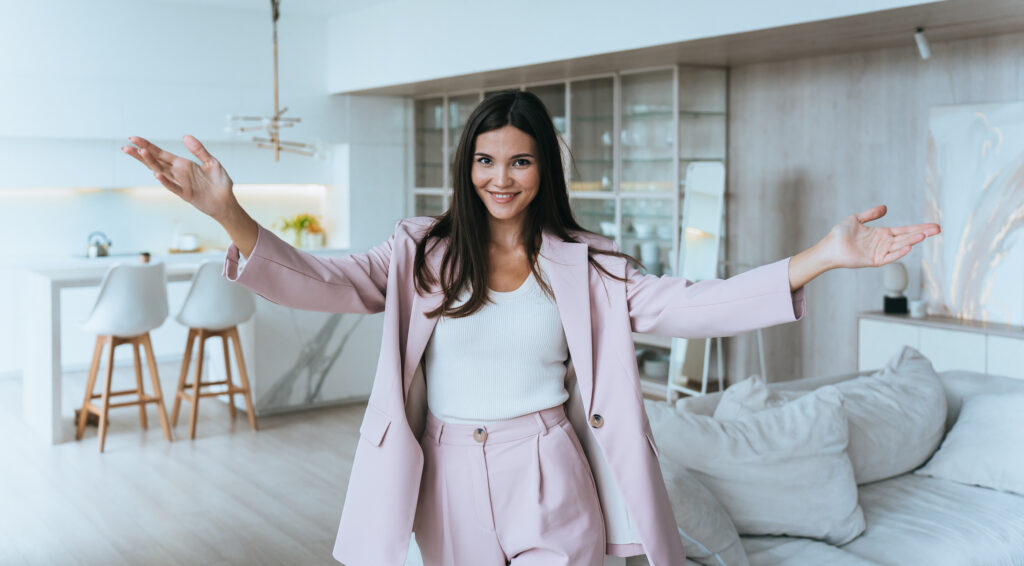 Smiling real estate agent in a light pink suit confidently presenting a modern, upscale home interior with open arms.
