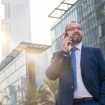 Confident businessman in a blue suit talking on a mobile phone, standing in front of modern glass buildings in Dubai with the Emirates Towers in the background.