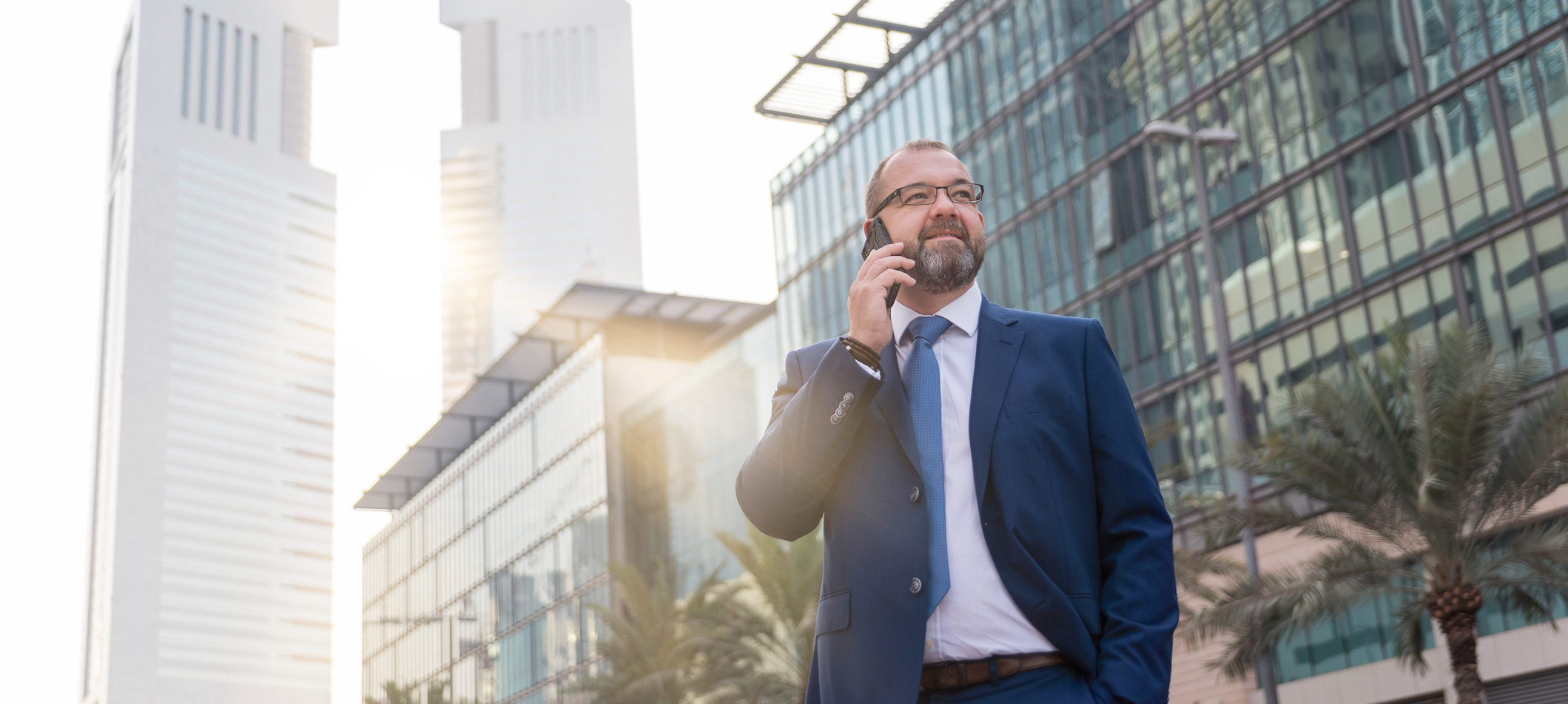 Confident businessman in a blue suit talking on a mobile phone, standing in front of modern glass buildings in Dubai with the Emirates Towers in the background.