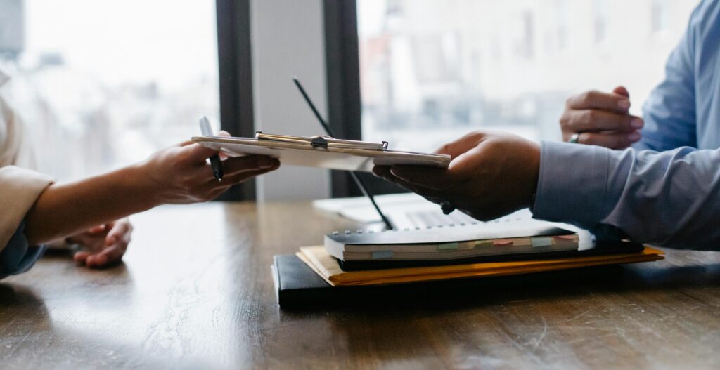 Close-up of two people exchanging a clipboard with documents across a desk, symbolising professional collaboration, review, or agreement in a modern office setting.