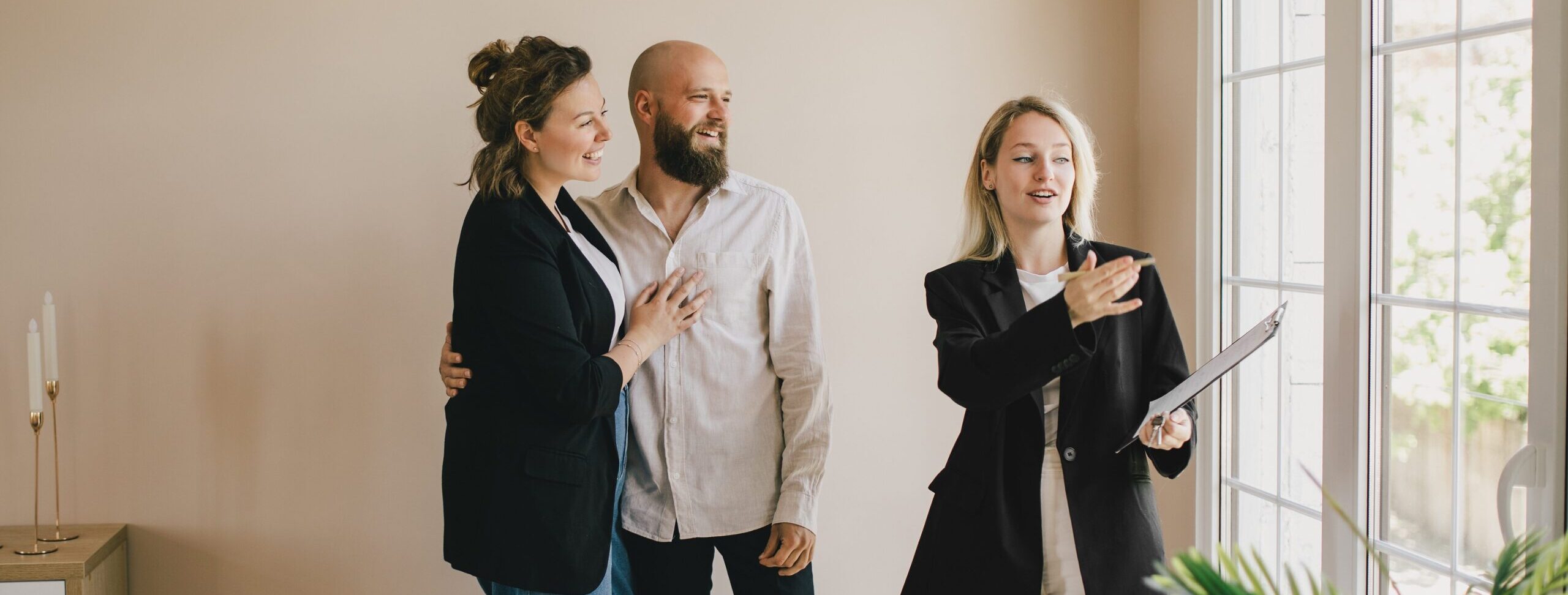 Smiling real estate agent showing a cheerful couple around a bright, modern home, gesturing toward large windows during a property tour.