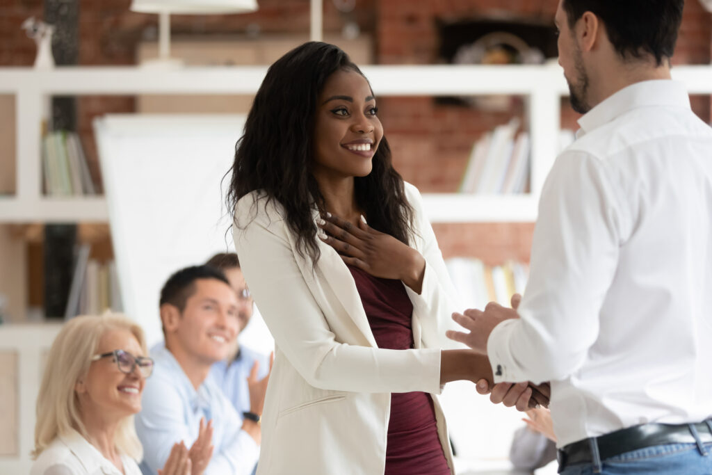 Smiling female real-estate agent receiving public recognition and a handshake from a colleague while the team applauds in the background.