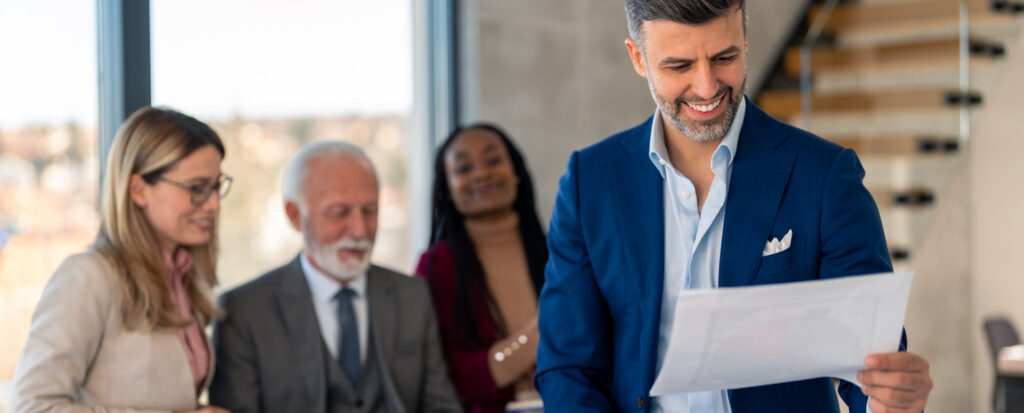 Real-estate team in a modern office—smiling agency leader in a blue suit reviews a document while three colleagues look on approvingly in the background.