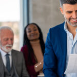 Real-estate team in a modern office—smiling agency leader in a blue suit reviews a document while three colleagues look on approvingly in the background.