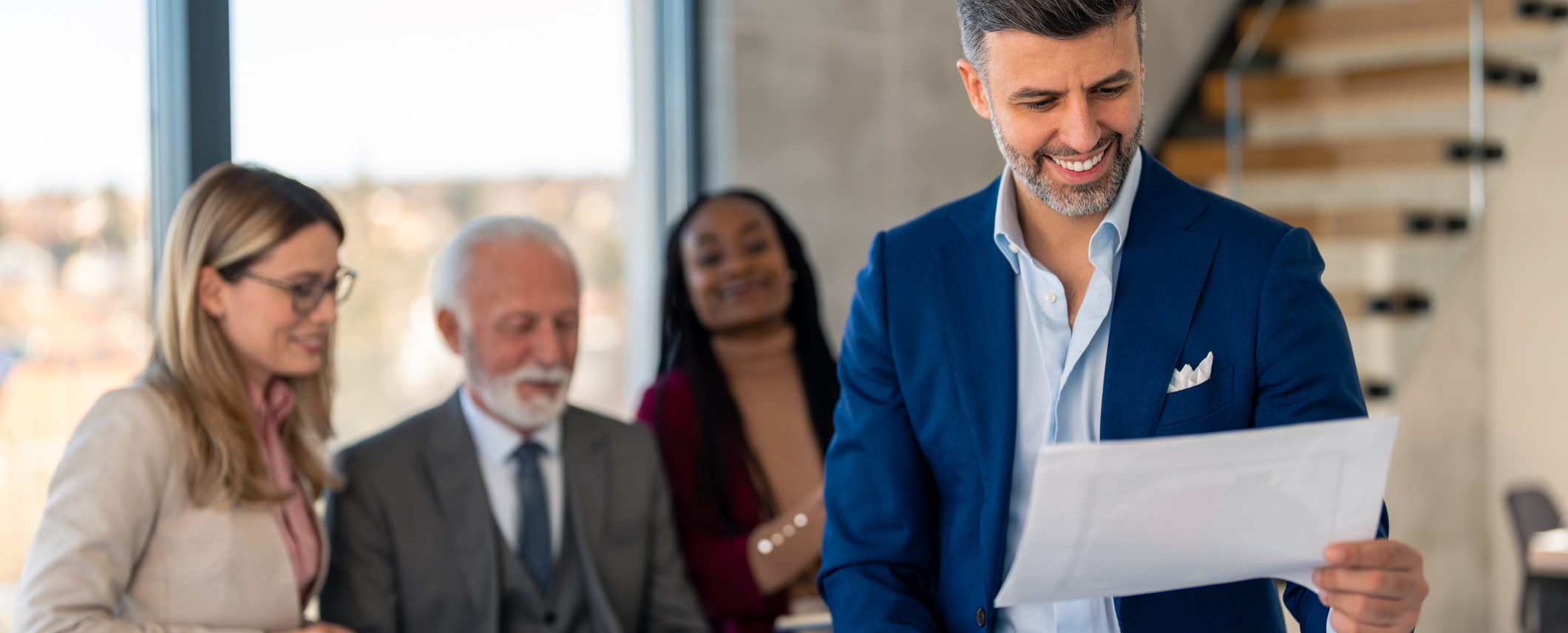 Real-estate team in a modern office—smiling agency leader in a blue suit reviews a document while three colleagues look on approvingly in the background.