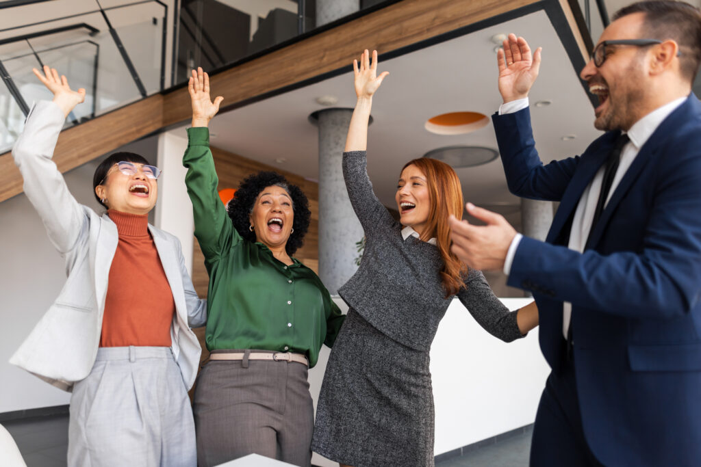 Diverse office team cheering with raised arms and bright smiles, celebrating a shared achievement in a modern workspace.