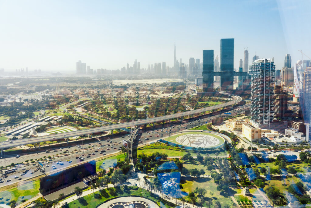 Aerial view of Sheikh Zayed Road cutting through green neighbourhoods towards Dubai’s skyline, with Burj Khalifa rising in the hazy distance.