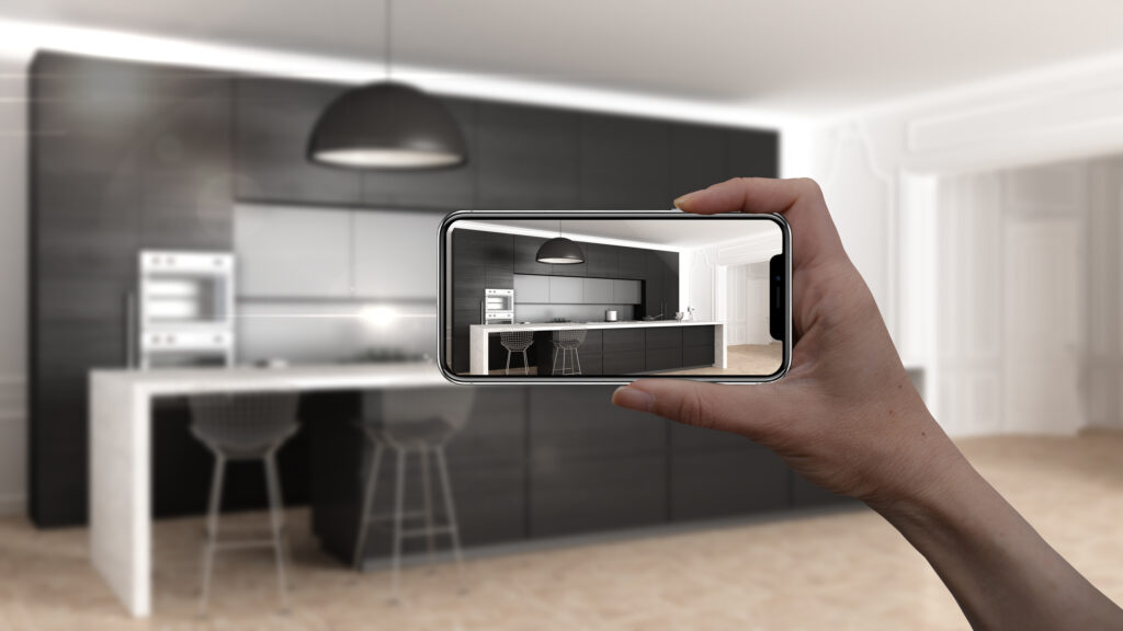 Hand holding a smartphone displaying a virtual rendering of a modern kitchen, superimposed over the real-world unfinished kitchen, illustrating the use of augmented reality in real estate virtual tours.