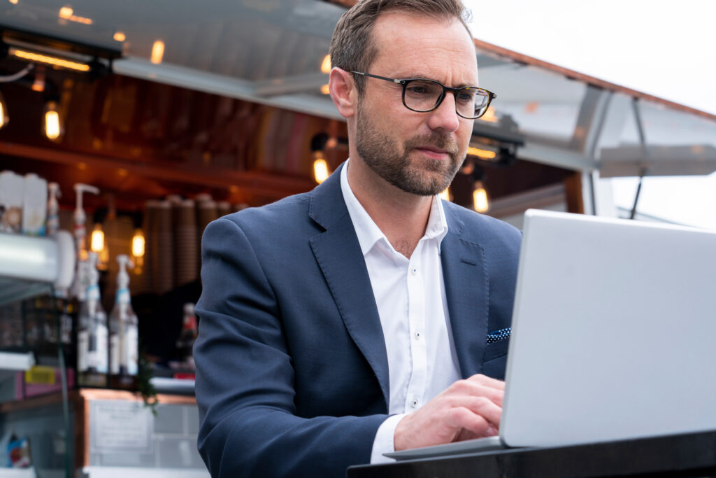 Future real estate agency owner in a navy suit and glasses typing on a laptop at an outdoor coffee stand, working on registering his business trade name in Dubai.