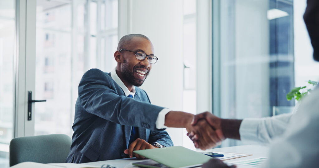 Smiling real estate professional in a business suit shaking hands across a desk during a welcoming or onboarding meeting in a bright, modern office.