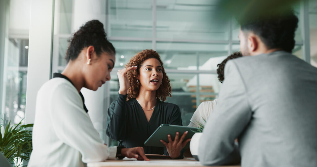 A diverse group of real estate professionals engaged in a discussion around a table in a modern office, with a confident woman at the centre gesturing while holding a tablet—symbolising collaboration and a strong team culture.