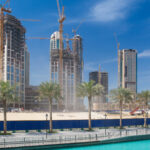 Dubai off-plan real estate development with high-rise towers under construction along a waterfront, showing cranes, palm-lined promenade, and active building progress
