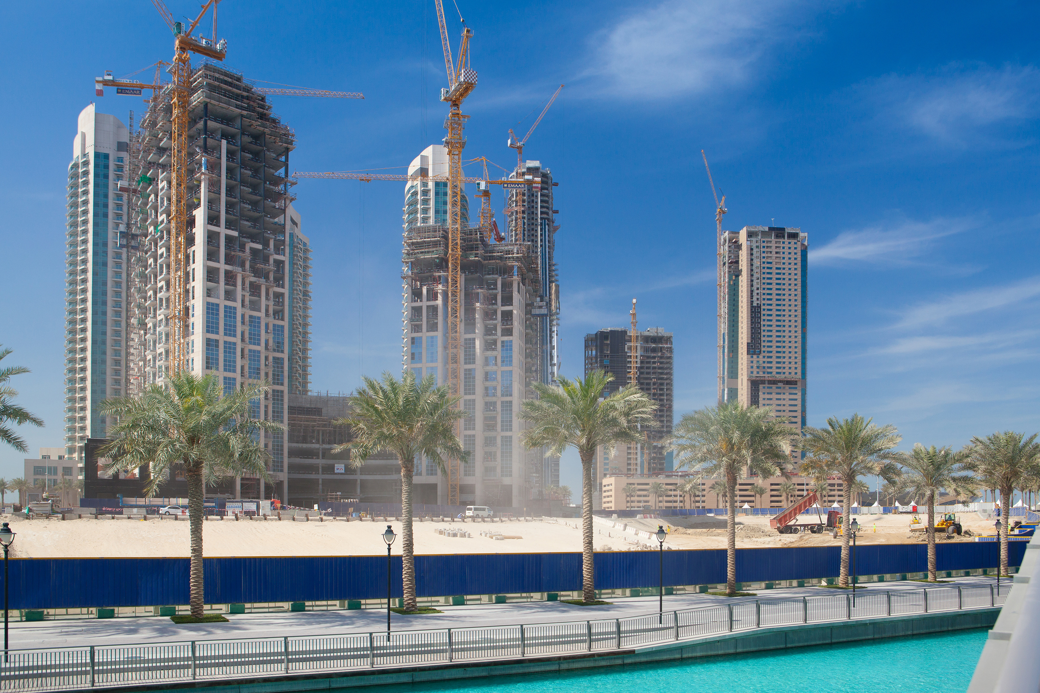 Dubai off-plan real estate development with high-rise towers under construction along a waterfront, showing cranes, palm-lined promenade, and active building progress