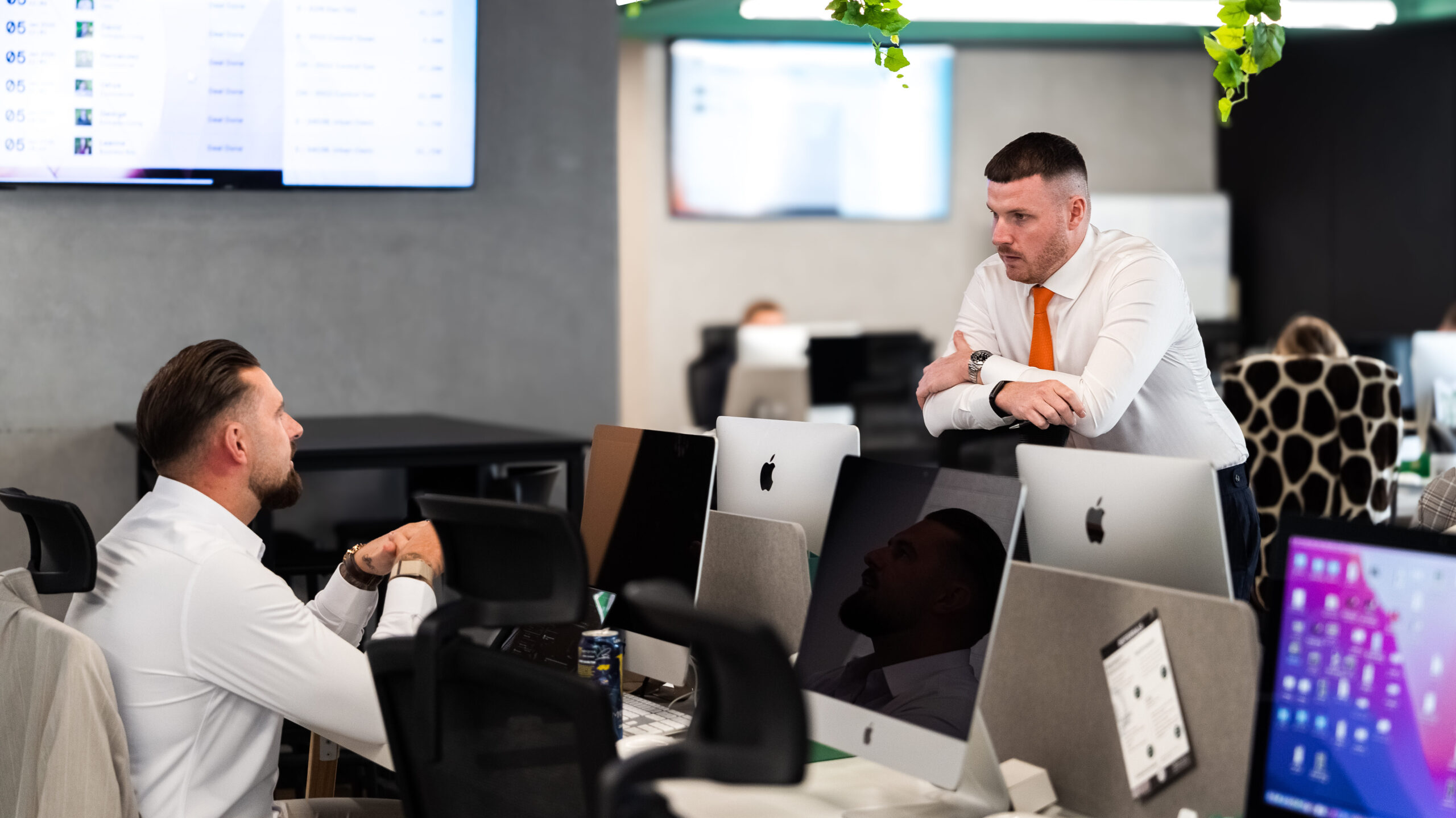 Real estate sales manager coaching a broker on the sales floor in a Dubai agency, reinforcing hands-on sales training, deal skills, and performance coaching beyond CRM systems and processes.